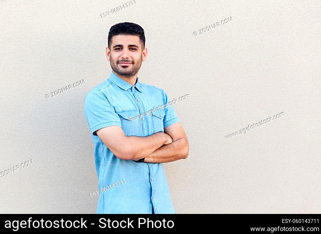 Portrait of confident successful handsome young bearded businessman in blue shirt standing, crossed arms and looking at camera with smile