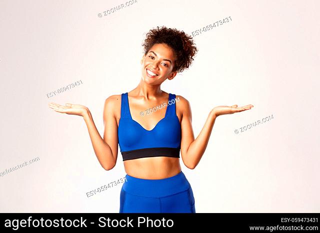 Waist-up shot of smiling african-american fitness girl in blue sport oufit, raising hands sideways, holding something or demonstrating
