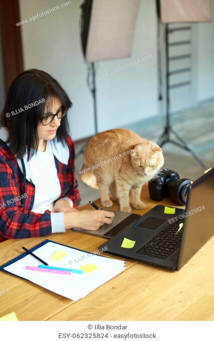 Creative female photographer with cute cat, using graphic drawing tablet and stylus pen, working at desk and retouch photo on tablet computer