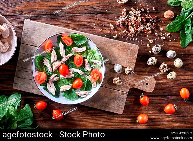 Healthy freshly prepared salad of quail eggs, meat, tomatoes and spinach in a plate on a wooden board on the kitchen table. Dietary lunch. Flat lay