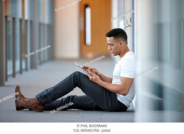 Young businessman sitting in office corridor using digital tablet