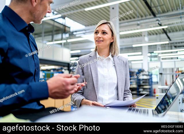 Businessman and businesswoman with laptop and documents talking in factory