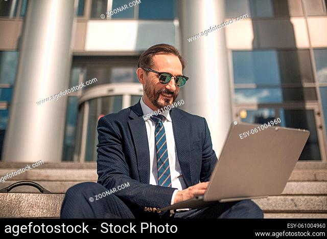 Successful businessman works on laptop sitting on steps of business center. Smiling man wearing business suit and sunglasses. High quality photo