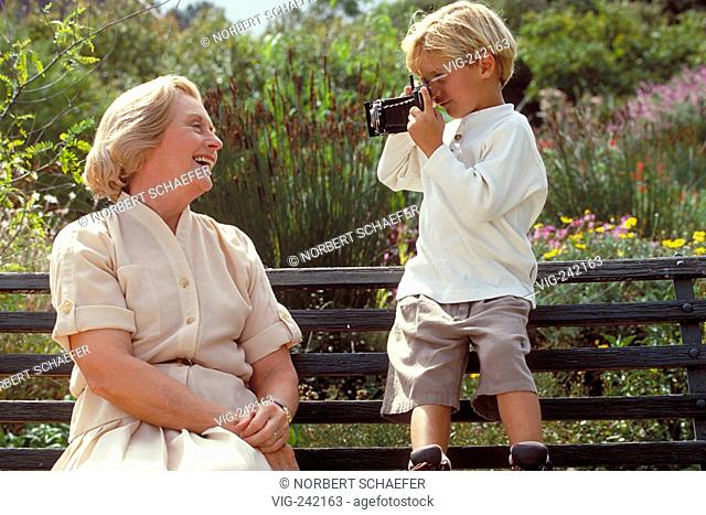 outdoor, blond 6-year-old boy stands with an old camera on a parkbench making a picture of his grandmother wearing a bright blouse and skirt  - GERMANY