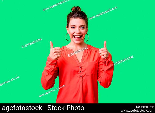 Thumbs up. Portrait of satisfied positive young woman with bun hairstyle, big earrings and in red blouse standing smiling widely, showing like gesture
