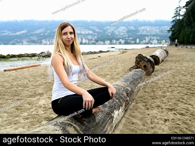 Young woman at the sandy beach of Stanley Park sitting on log, her arms are down and doing some acrobatic exercise movement, Vancouver, British Columbia, Canada