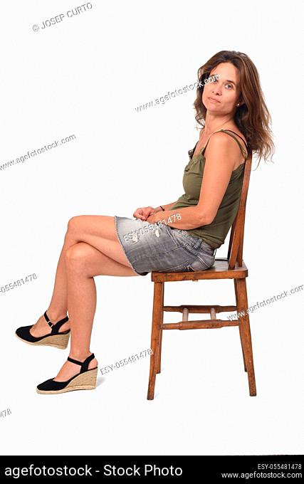 side view of a serious woman in denim skirt sitting on a chair and looking at camera on white background, legs crossed