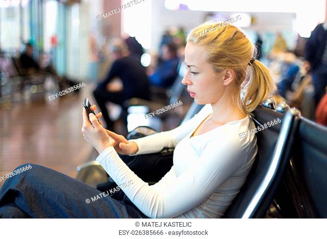 Casual blond young woman using her cell phone while waiting to board a plane at the departure gates. Wireless network hotspot enabling people to access internet...
