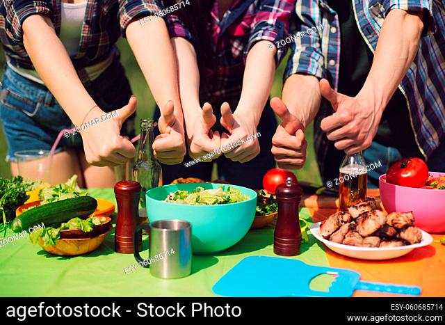 Group of friends showing thumbs up to table of food. Group of young peope showing thumbs up signs with both hands to big table of picnic food they have prepared