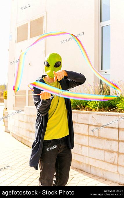 Man wearing alien mask playing rainbow rhythmic ribbon on footpath
