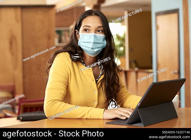 Portrait of young ethnic woman wearing yellow sweater with black and white striped blouse and face mask, sitting at bar in kitchen of downtown loft with iPad