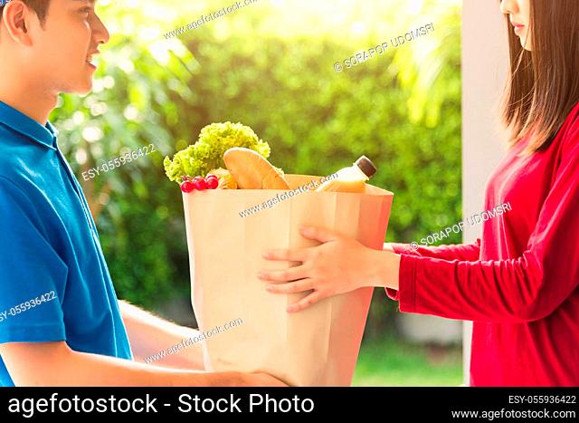 Asian young delivery man in uniform making grocery fast service giving fresh food in paper bag to woman customer receiving at house door under pandemic...