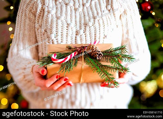 Woman holding ecological Christmas present on the background of a Christmas tree. The gift is wrapped in craft-paper with a spruce branch
