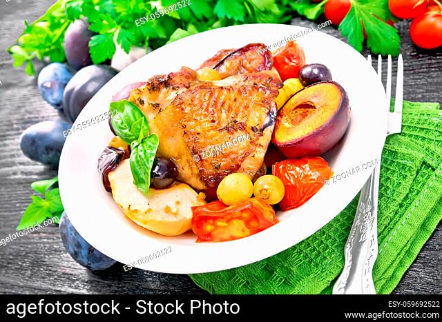 Baked chicken with tomatoes, apples, plums and grapes in a plate on a napkin, garlic, parsley and basil on wooden board background