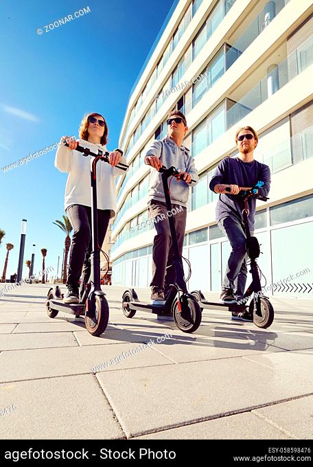 Three best friends young 20s -30s girl and guys spend time outdoors gathered together driving on electric scooter modern land vehicle, easy comfort usage