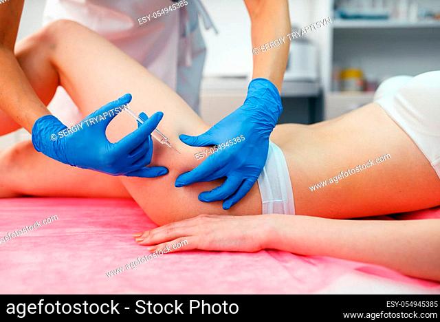 Cosmetician in gloves gives botox injection in the thigh to female patient on treatment table. Rejuvenation procedure in beautician salon