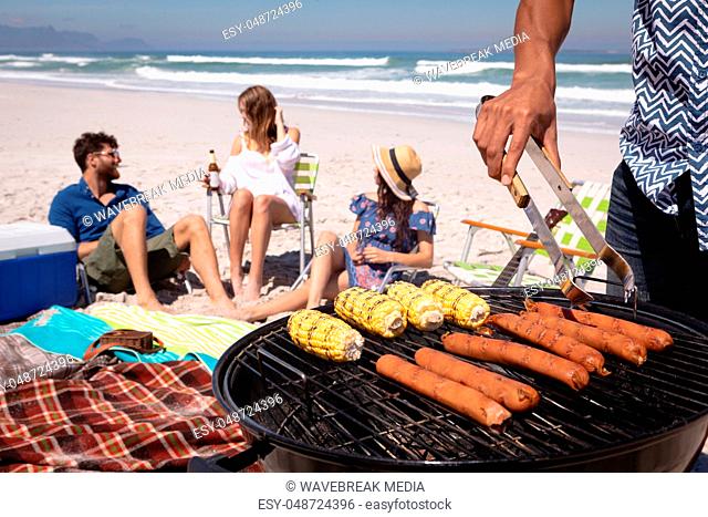 Mid-section of a man doing a barbecue while his friends in background at beach