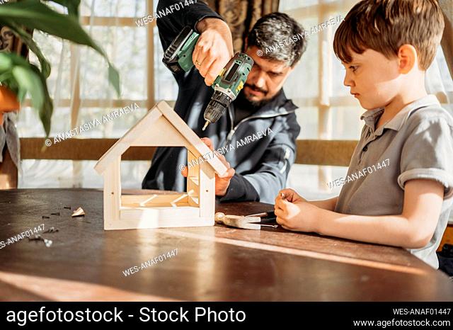 Father drilling birdhouse on table with son at home