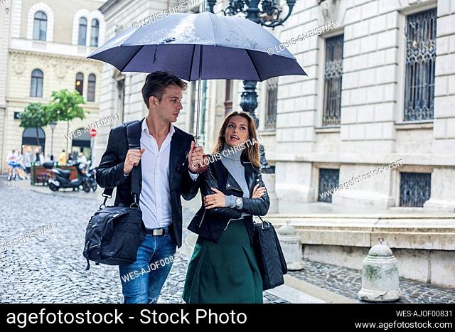 Businesswoman with male colleague walking under umbrella on street in city