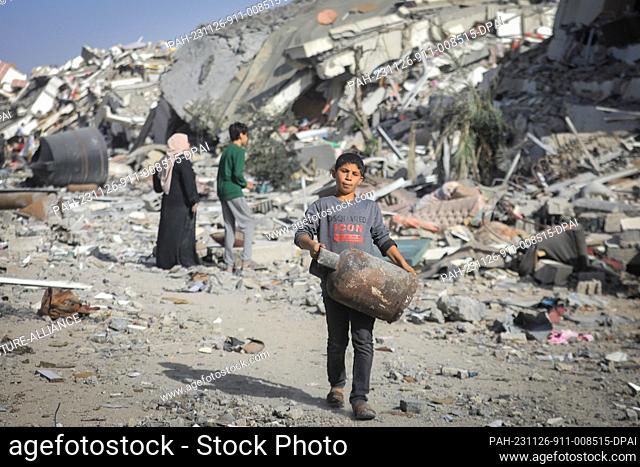 26 November 2023, Palestinian Territories, Gaza: A Palestinian child carries household items as he walks around destroyed buildings in Al-Zahra City