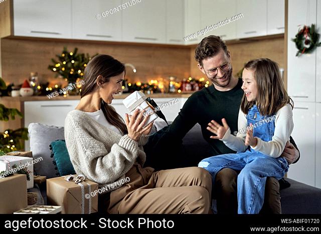 Happy woman showing gift to daughter sitting on father's lap at home