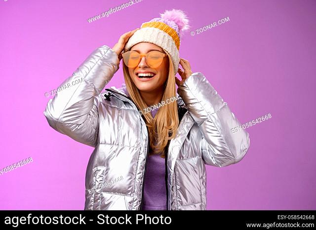 Carefree amused charming blond girlfriend having fun enjoying awesome sunny winter day ski resort vacation wearing sunglasses silver stylish jacket put on hat...