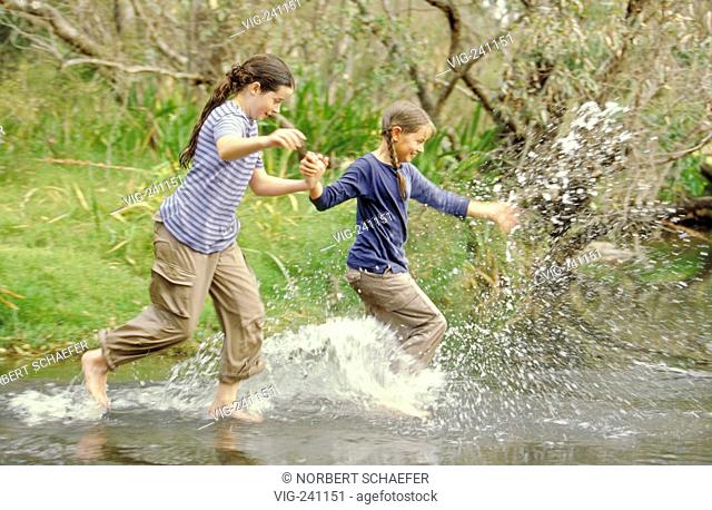 portrait, 2 girls, 12 years, with long hair, wearing t-shirt and trousers running bare feeted through a rivulet  - GERMANY, 25/01/2004