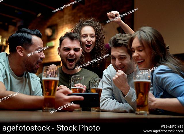 Group of cheerful soccer fans having beer and watching a match on smartphone in a pub