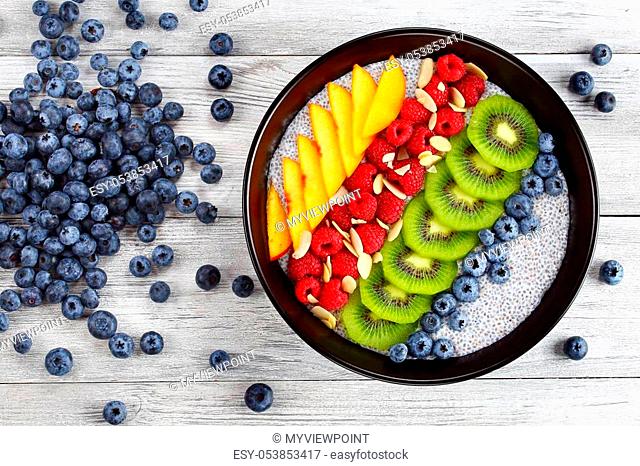 delicious chia seeds pudding with raspberry, blueberry, peach and kiwi fruit slices in black bowl on old wooden table, with pile of blueberries on background