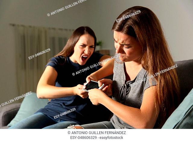 Two friends or sisters fighting for a smart phone sitting on a couch in the living room at home