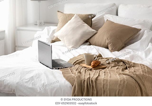 laptop, cup of coffee and croissant on bed at home