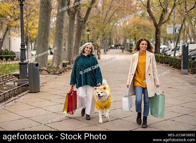 Happy senior woman holding shopping bags walking with granddaughter and dog at footpath