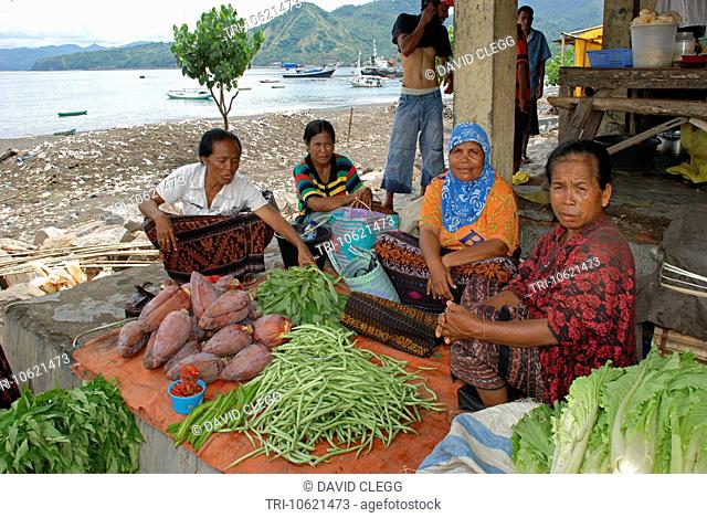 Four women sit on a concrete step at Ende market with vegetable laid out on plastic sheet with a view of the harbour with shipping a dirty sandy beach and green...