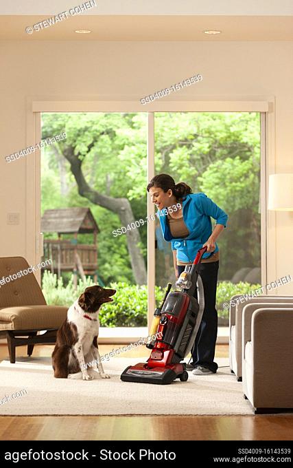 woman at home using vacuum on rug in living room with her pet dog looking on. view of back yard through double sliding door