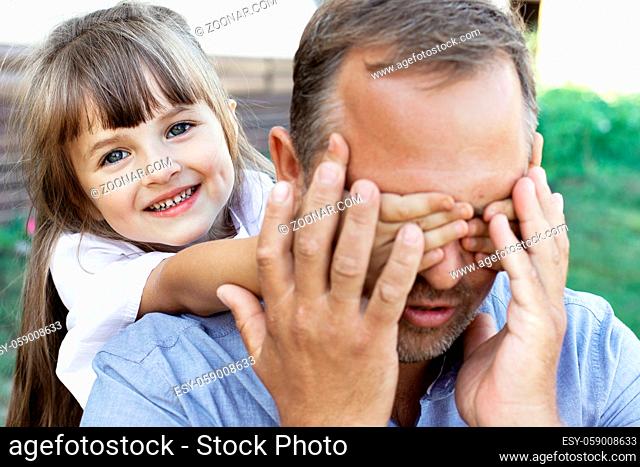 Little beautiful girl playing with her father in nature. A cheerful child covers the parent's eyes with his hands. Family happiness to be together
