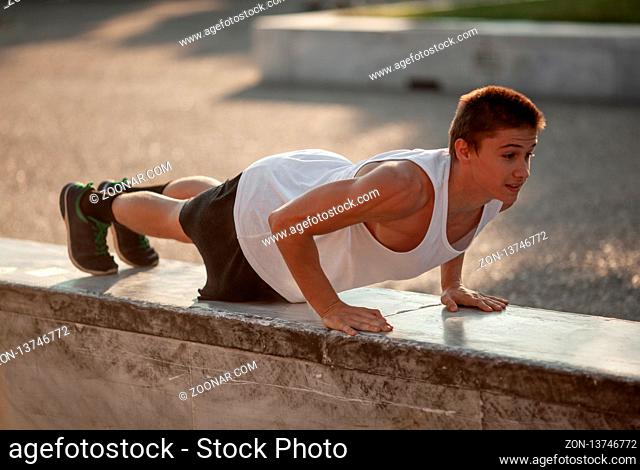A teenage boy makes outdoor press-ups, standing on a wide stone fence
