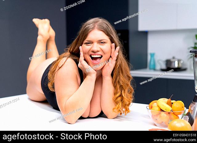 Overweight girl with big breasts laying on the kitchen table. An obese young sexy chubby white girl in black swimsuit at modern kitchen with fruits next to her