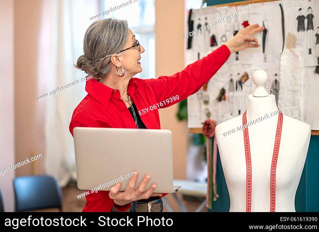 Good idea. Profile of a gray-haired woman in glasses holding laptop smiling touching sketch hanging on wall in atelier