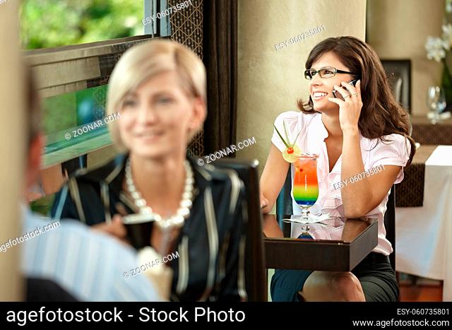 Young businesswoman sitting at table in cafe, talking on mobile phone, drinking cocktail