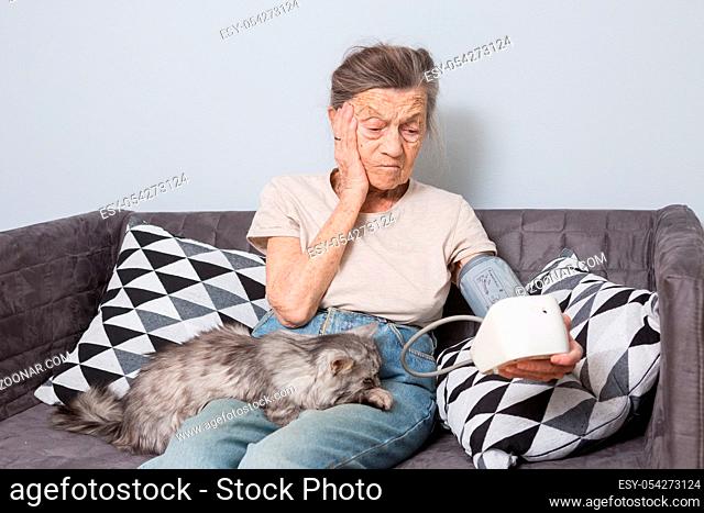The topic is very old person and health problems. A senior Caucasian woman, 90 years old, with wrinkles and gray hair, sits home on sofa with pet cat and uses...
