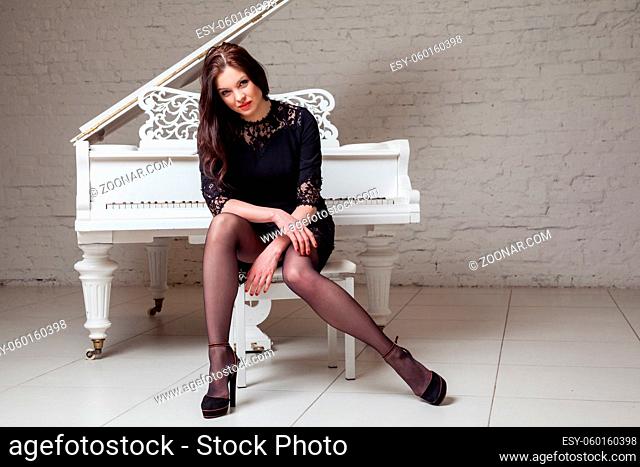 Sensual brunette woman in lace black classic dress sitting near white piano and looking at camera. indoor studio shot on white brick wall