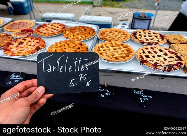 A person's hand is seen closeup holding a small French price tag, saying tartlets, by a display of freshly baked sweet tarts on a bakery stand during a market