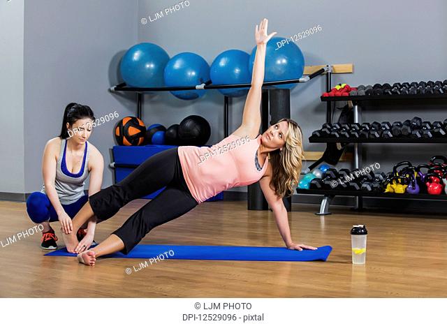 A middle-aged women doing a side plank yoga exercise at the gym with her personal trainer giving assistance; Spruce Grove, Alberta, Canada