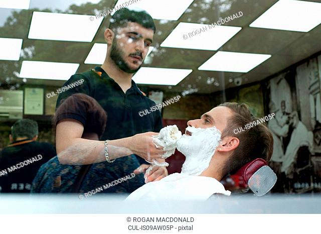View through window of young man in barbershop applying shaving cream to customers face