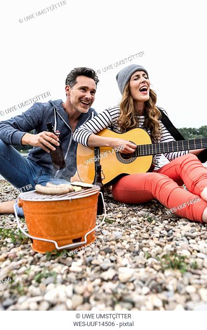 Happy couple with beer bottle, guitar and grill relaxing on pebble beach