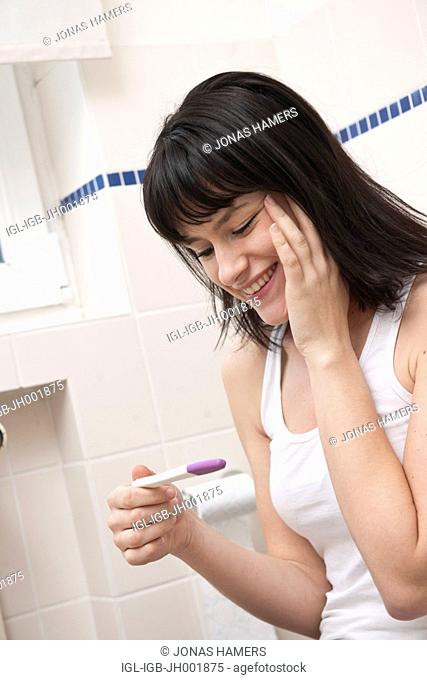 This picture shows a young caucasian woman with brown hair smiling as she watches a pregnancy test