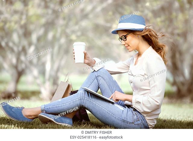 Happy young woman a disposable coffee cup sitting on the grass and using touch pad on her tablet in a summer park