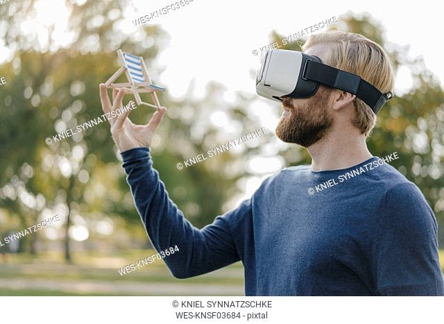 Man in autumnal park wearing Virtual Reality Glasses looking at miniature beach lounger