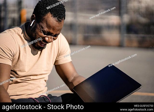 Man laughing while sitting with digital tablet on sports court during sunny day
