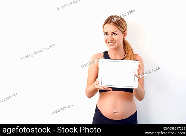 Smiling friendly young pregnant woman holding up a blank white modern tablet with screen to the viewer over a studio background with copy space